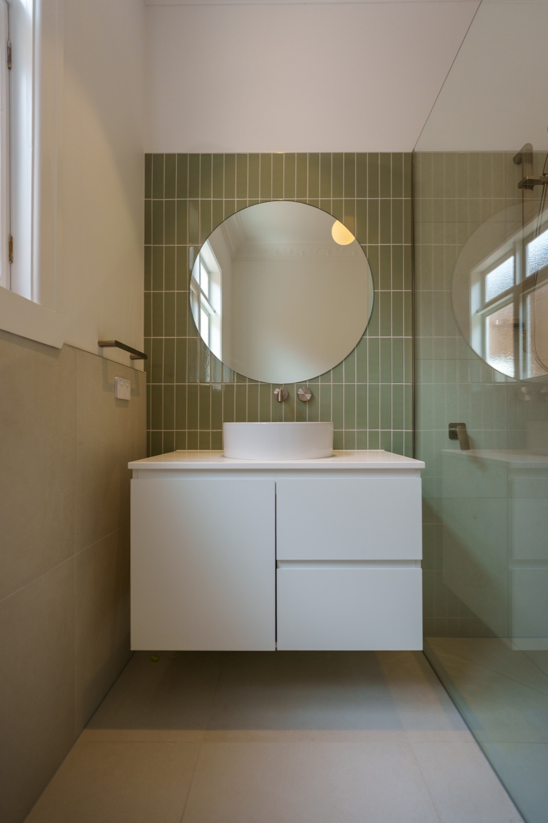 A bathroom in green square tile with a round mirror and floating vanity