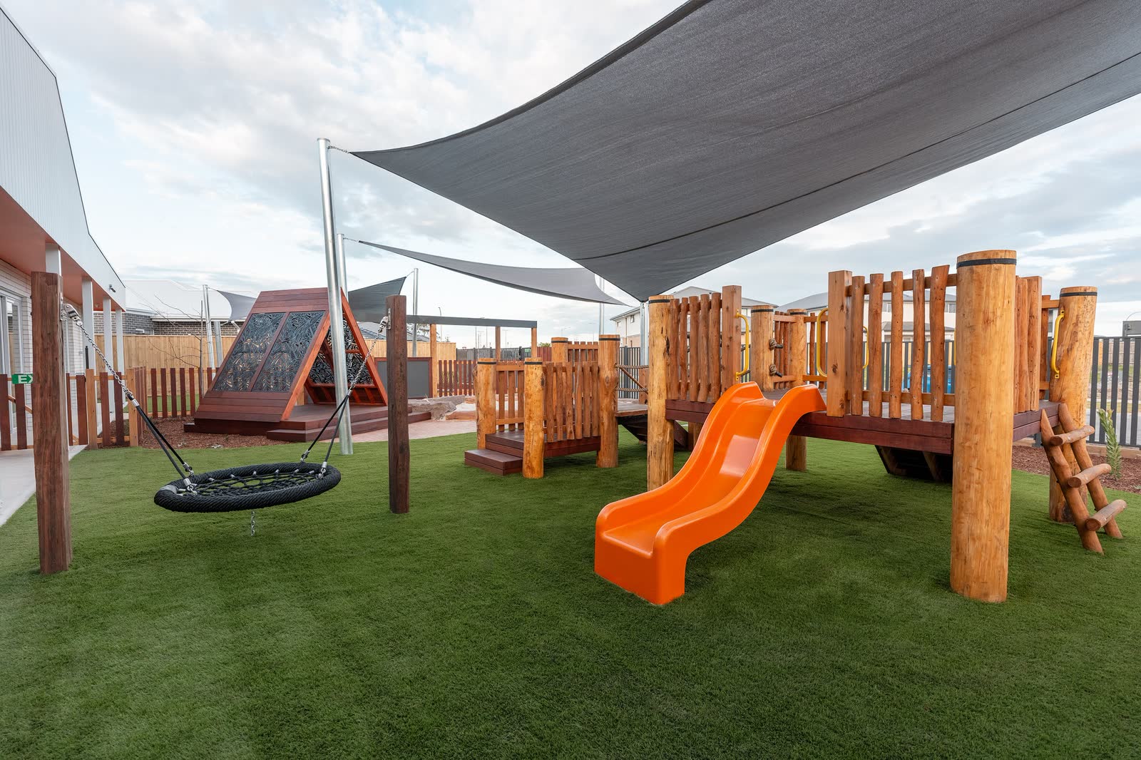 A timber play structure with an orange slide beneath a shade sail