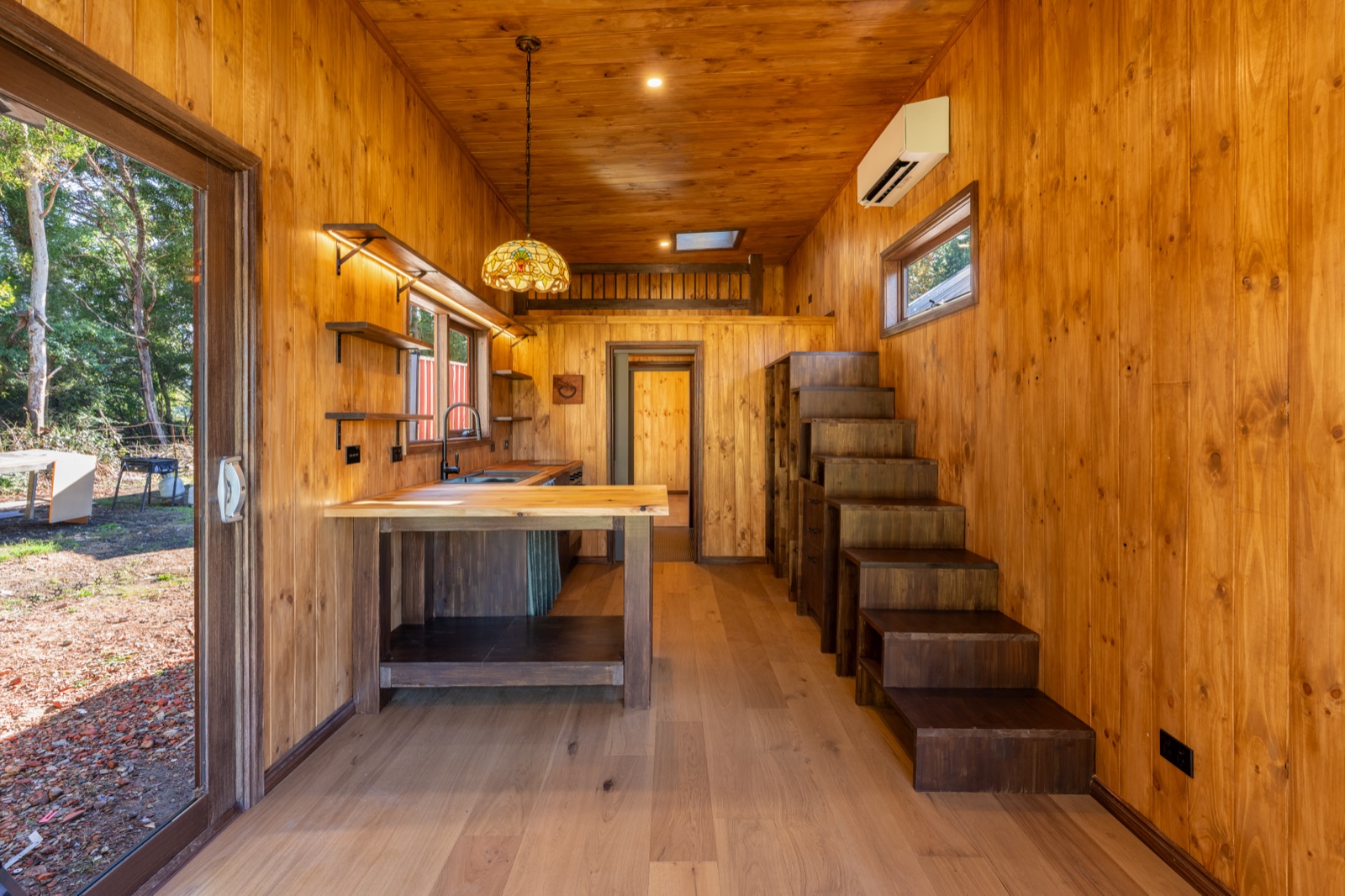 A timber lined tiny home interior with a kitchen island and stairs to the loft