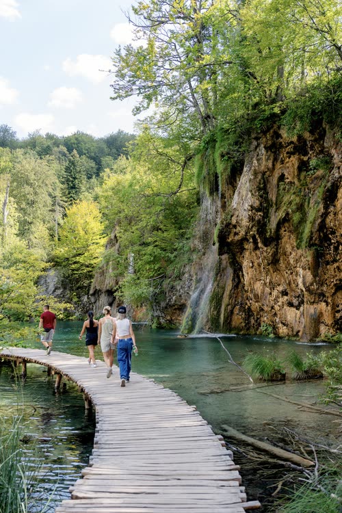 Walkers on the boardwalk beneath a mossy waterfall cliff, Croatia