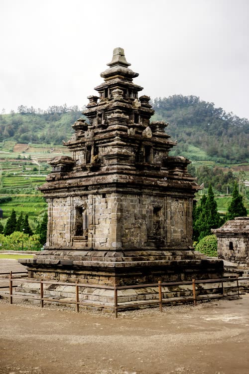 A small stone Hindu temple standing against terraced hills in central Java