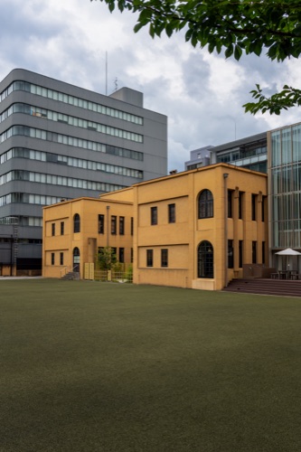 A yellow rendered arts building beside a glass tower under an overcast sky