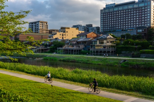 Cyclists on the Kamo river path at dusk with the hotels of the far bank behind