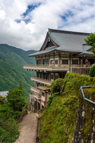 A temple hall perched on a steep green hillside with a storm sky above