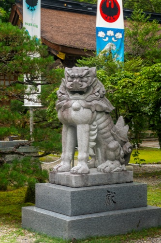 A stone komainu guardian lion before a shrine hung with banners and pines