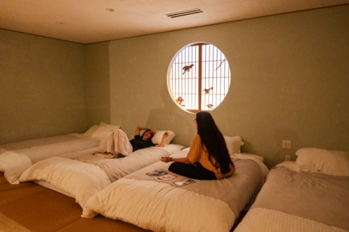Two travellers on futons in a tatami room with a round window and hanging paper birds