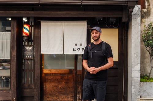 A man in a cap standing before a barber shop doorway with a white noren