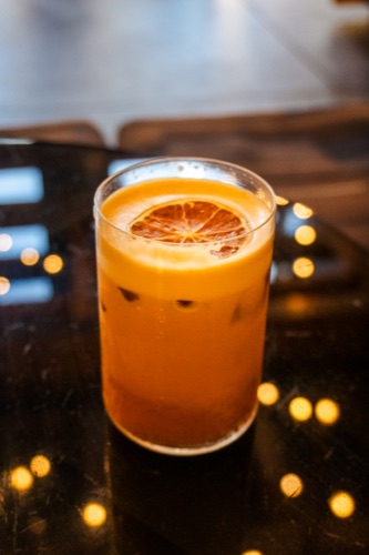 A glass of iced orange juice with a dried citrus slice on a black bar top with bokeh lights