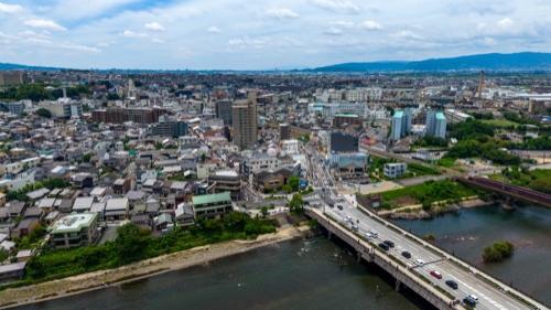An aerial view across the rooftops of a Japanese city to the river bridges and the hills beyond