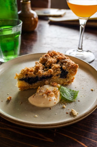 A slice of blueberry crumble cake with cream and mint on a stoneware plate