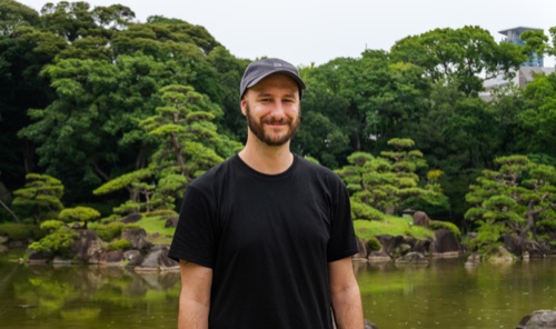 A man in a black tee and cap standing before a pond and clipped pines in a garden