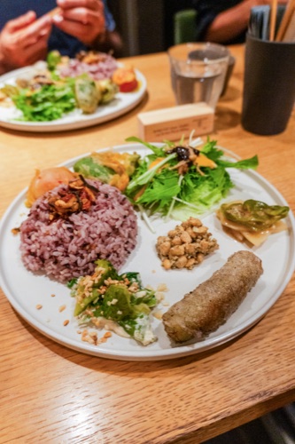 A plate of purple rice, greens, tempura and a croquette at a wooden table