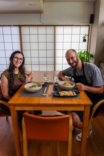 Two people in aprons sitting down to bowls of noodles and gyoza at a timber table
