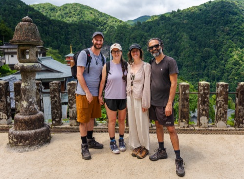 Four hikers at a stone balustrade with a lantern and forested mountains behind