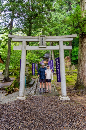 Two hikers in dark gear standing beneath a stone torii at a forest shrine