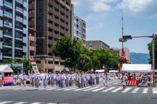 A procession of festival participants in grey crossing a wide Kyoto intersection by a tall float