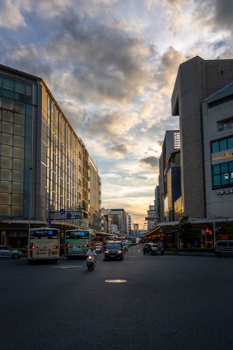 A wide Kyoto street at sunset with buses and scooters under a dramatic sky
