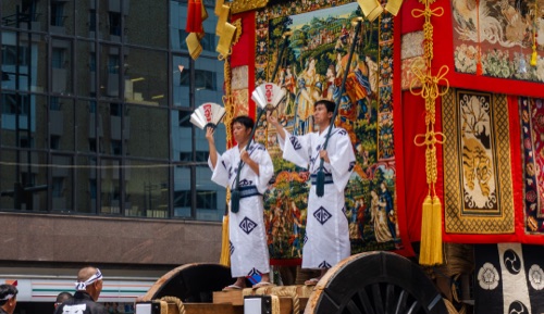 Two men in white festival dress waving fans from the front of an ornate tapestry covered float
