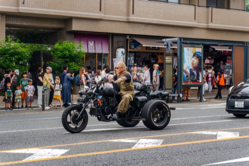 A man on a black trike motorcycle riding past a crowded bus stop in Kyoto