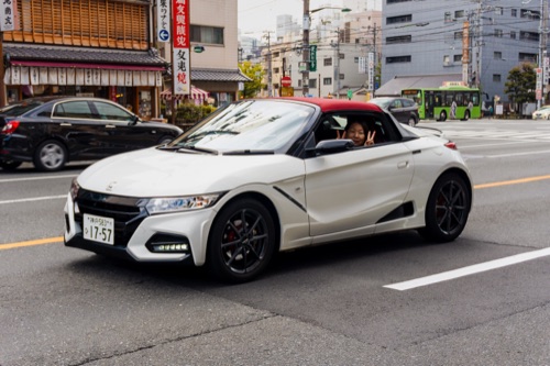 A white Honda S660 roadster passing on a Kyoto street, the driver waving