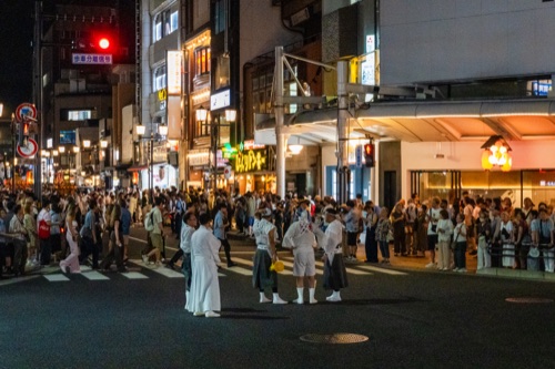 Festival staff in white crossing a neon lit intersection at night with crowds behind
