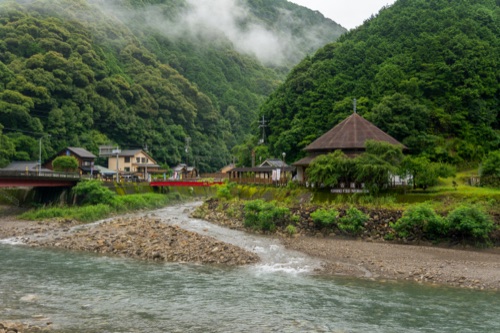 A river meeting a gravel bar below a village roof and mist on the forested hills