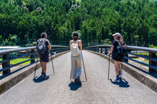 Three hikers with trekking poles crossing a bridge towards forested hills