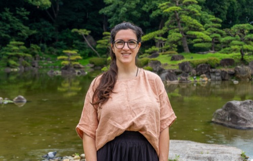A woman in a pink linen top standing before a pond and clipped pines in a garden