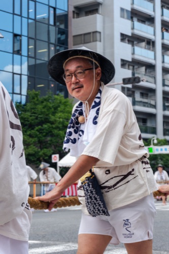 A man in a straw hat and white festival coat hauling a rope through the city street
