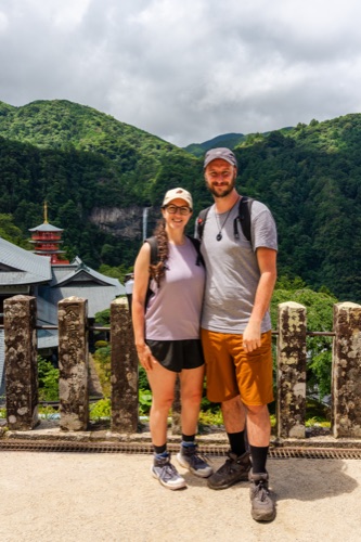 A couple in hiking gear at a stone railing with a pagoda roof behind them