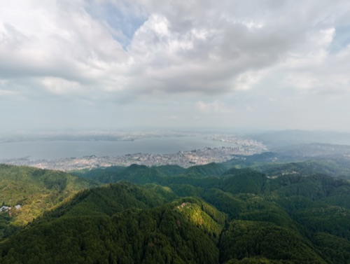Forested ridgelines dropping to a city on the shore of a lake, shot from altitude in soft haze