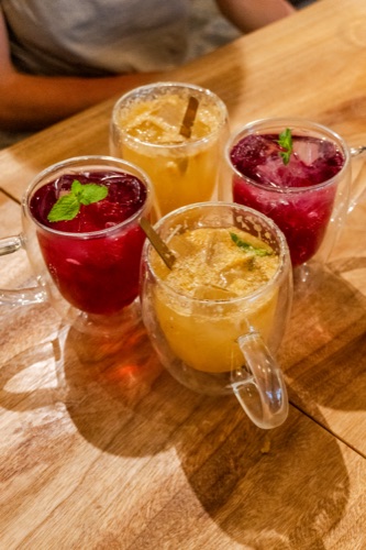 Four iced drinks in double walled glasses, two berry red and two citrus yellow, on a timber table