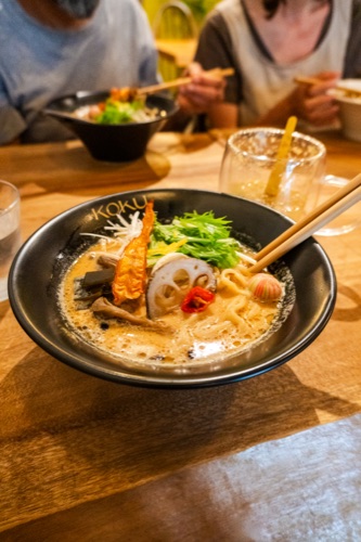 A bowl of ramen with lotus root, mushroom and greens in a creamy broth, friends eating behind