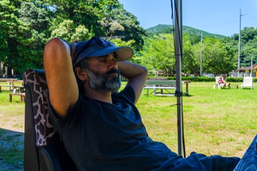 A bearded man in a cap resting with hands behind his head at a riverside campsite