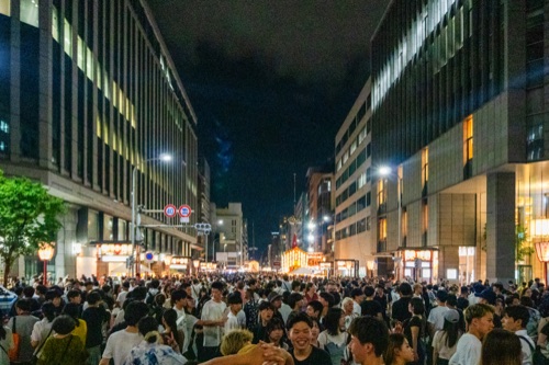 A Kyoto street packed with festival goers at night between lit office towers