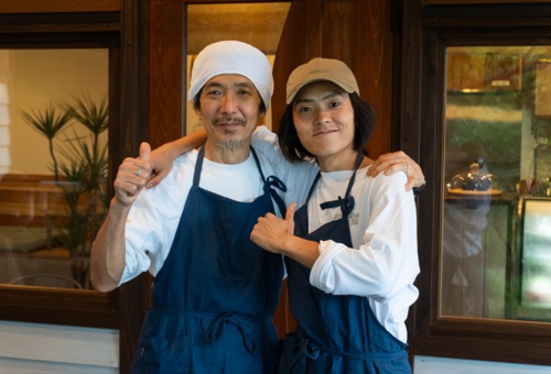 Two cooks in navy aprons with arms around each other at a restaurant doorway