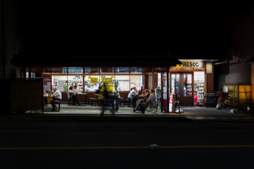 A convenience store glowing on a dark street, people on the benches outside