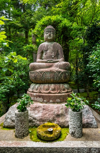 A stone seated Buddha on a lotus base framed by green, with stone vases of leaves