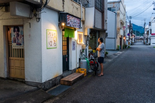 A quiet lane of shuttered shopfronts at dusk with a man standing outside a lit cafe