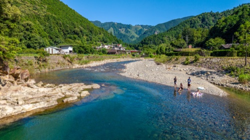 A clear turquoise river bending past a stone beach with a few swimmers, forested hills behind