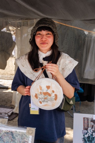 A woman in a crocheted hat holding an embroidered hoop at a craft market stall