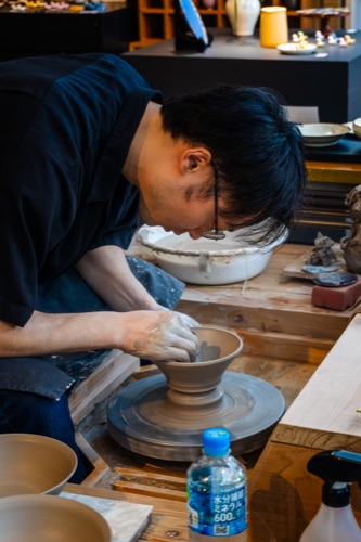 A potter bent over a wheel throwing a bowl in a small studio
