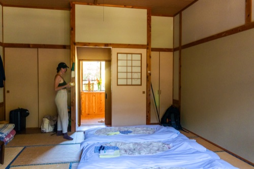 A traveller in a tatami guesthouse room with futons laid out and a sunlit doorway