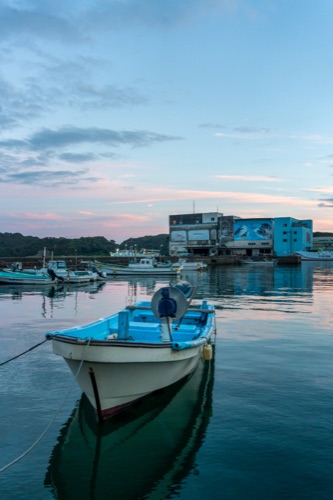 A small fishing boat moored in a calm harbour at dusk with a mural painted warehouse behind
