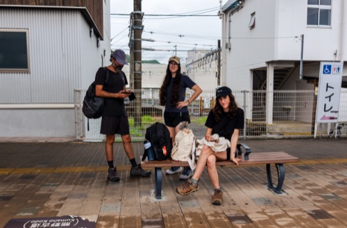 Three hikers with packs waiting on a bench outside a small country train station