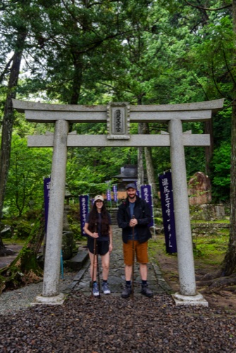 Two travellers beneath a stone torii on a gravel approach lined with purple banners