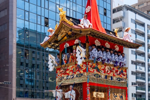 Musicians in blue and white seated on a tapestry hung festival float against glass office towers