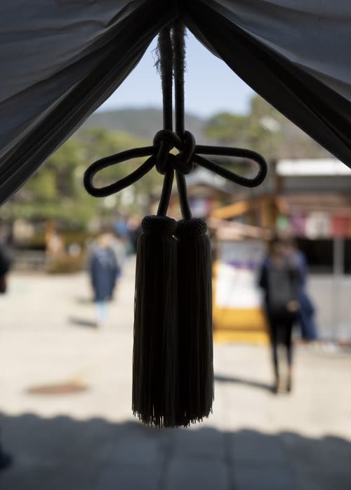 A heavy black silk tassel hanging from a temple canopy, visitors blurred beyond