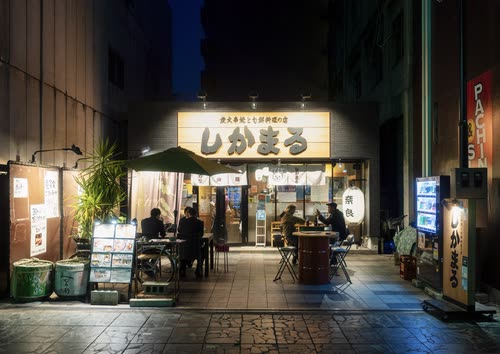 Diners at the counter of a small lit restaurant on a Japanese street at night