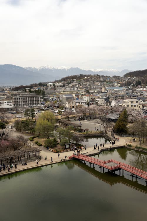 A red arched bridge over a park lake with the snow-capped Japanese Alps beyond